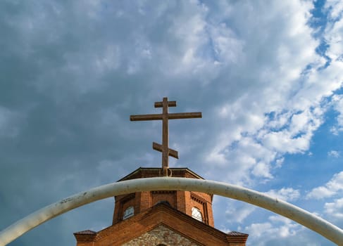 White semi-circular arch of iron pipe with dark Orthodox cross over it before the tower of the ancient temple of bricks against the blue sky with clouds.