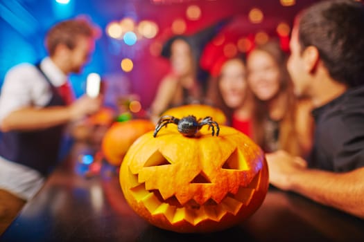 Carved pumpkin and spider on background of group of friends celebrating Halloween in bar
