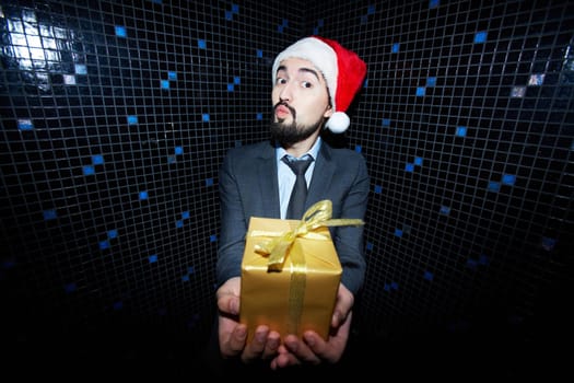 Bearded businessman in Santa cap holding giftbox and looking at camera in night club