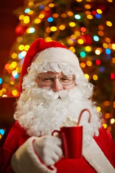 Santa Claus holding red cup with latte on background of sparkling xmas tree