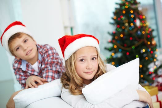 Adorable girl in Santa cap looking at camera with cute boy on background