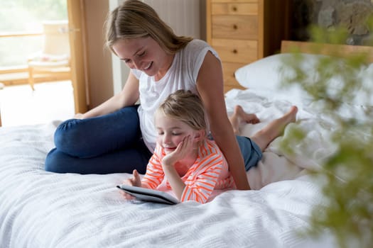 Mother and daughter lying on a bed at home together, using a digital tablet.