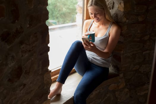 Woman sitting on her windowsill whilst using a smartphone and enjoying a hot drink.