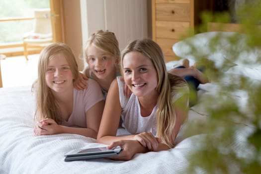 Sisters lying on a bed in their home with a digital tablet, smiling for the camera.