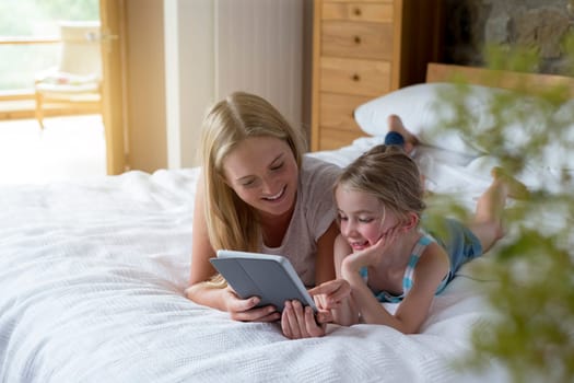 Mother and daughter lying on a bed at home together, using a digital tablet.