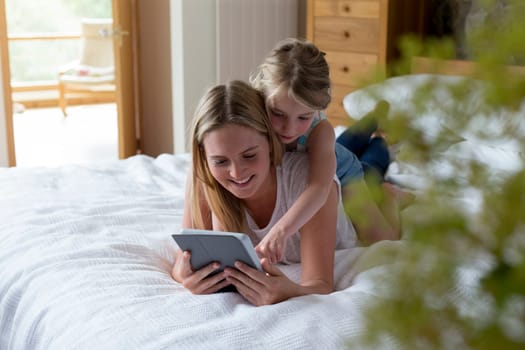 Mother and daughter lying on a bed at home together, using a digital tablet.