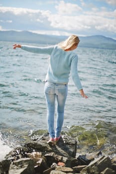 Rear view of blond girl in jeans and pullover standing on stone by the seaside