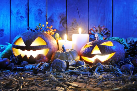 illuminated Halloween pumpkins,candles,nuts,corn cobs and apple on straw in front of old weathered wooden boards in the blue evening light