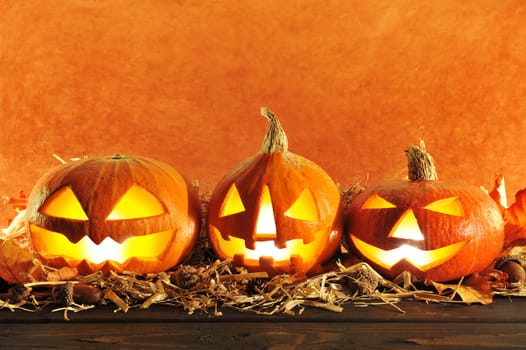 Three red illuminated Halloween pumpkins with straw on old weathered wooden boards against a brown background