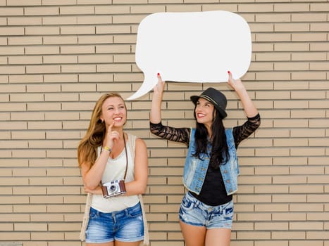 Two beautiful and young girlfriends holding a thought balloon, in front of a brick wall