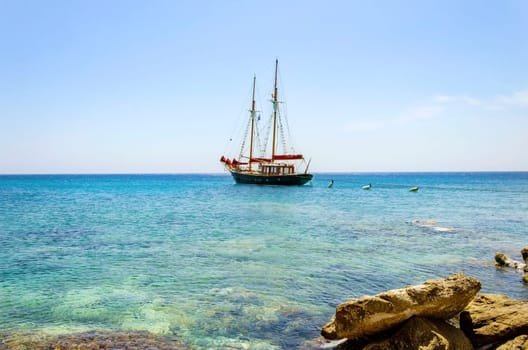 A traditional wooden sail boat anchored on the coast of Mykonos, Greece. The wind sails are down and it is tied on the rocks by a rope. A very typical greek island summer holiday scene on crystal clear blue sea.