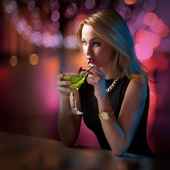 Young attractive woman looking around leaning on the bar counter while sipping her cocktail. Young lady in a club. Night life