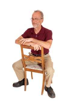 A serious older man in beige pants and burgundy t-shirt sitting backwards.on a chair, isolated for white background..