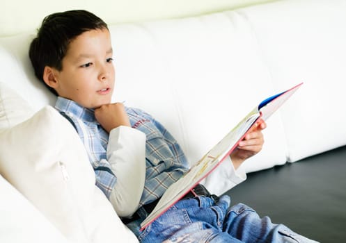 thoughtful little boy sitting on a sofa and reading a book