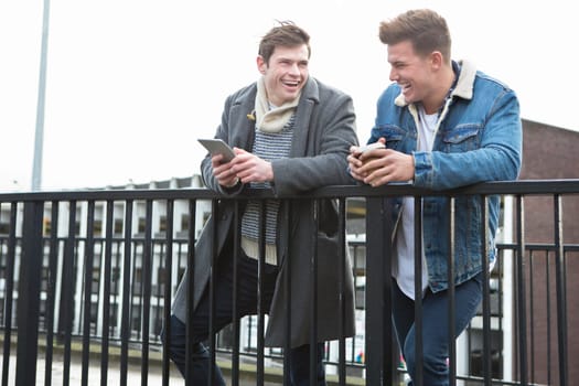 Two young men talking in the city. One is holding a smartphone, the other is holding a disposable coffee cup.