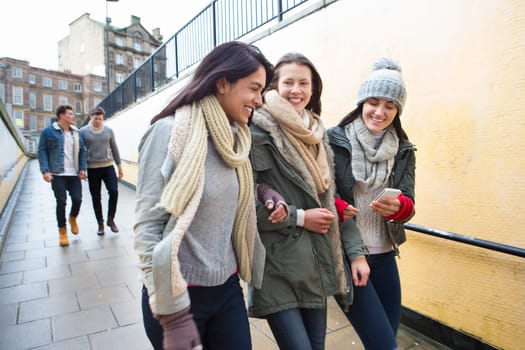 Three young women walking in the city, looking at something on a smartphone. Two men can be seen walking behind them.