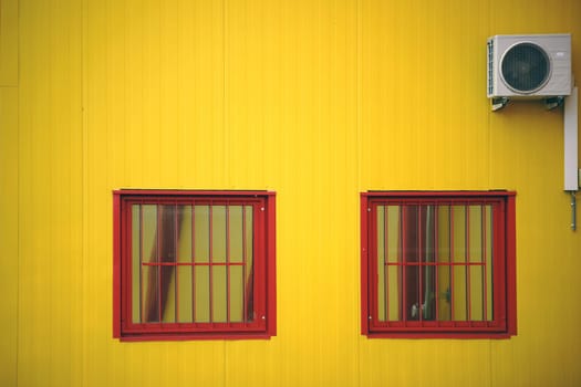 The yellow side wall of a shopping center with two barred windows and a fan.