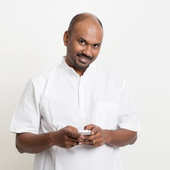 Portrait of mature casual business Indian man texting using smartphone, standing on plain background with shadow.
