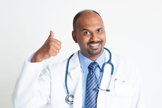Portrait of happy mature Indian male medical doctor in uniform showing thumb up, standing on plain background with shadow.