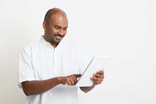 Portrait of mature casual business Indian man using digital tablet pc, standing on plain background with shadow.