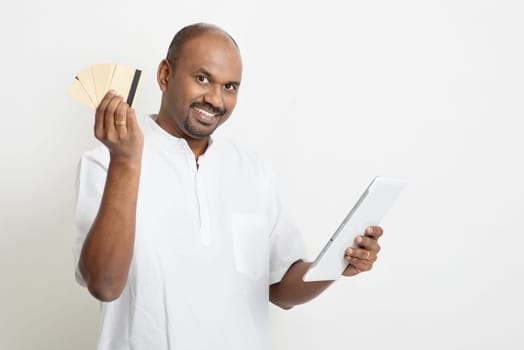 Portrait of mature casual business Indian man using digital tablet pc and credit card for online purchase, standing on plain background with shadow.