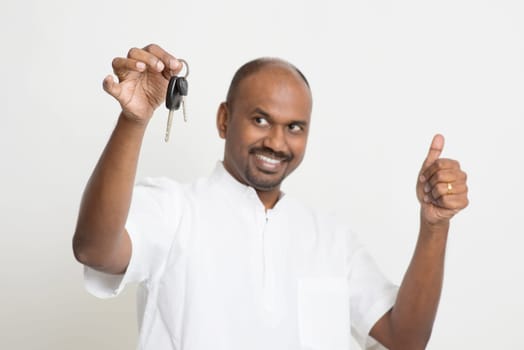 Portrait of Indian man holding new house key and thumb up, real estate property agent concept, focus on key, standing on plain background with shadow.