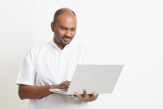 Portrait of mature casual business Indian man using laptop computer, standing on plain background with shadow.