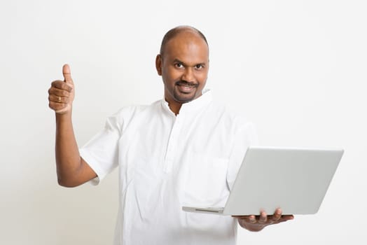 Portrait of mature casual business Indian man using laptop computer, looking at camera and thumb up, standing on plain background with shadow.