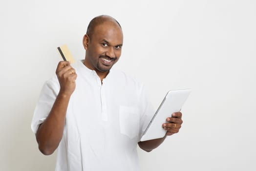 Portrait of mature Indian man shopping online using tablet pc, making payment by credit card. Asian man standing on plain background with shadow and copy space.