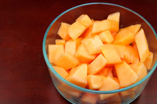 One orange cantaloupe melon that has been cut into pieces and placed into a in a clear, round glass bowl and placed on a medium dark brown wood surface.