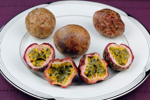 Three whole and four halves of a purple brown skinned passion fruit with yellow pulp  surrounding edible dark green seeds on a round white plate placed on a dark purple place mat.