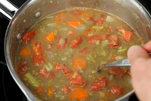 A hand of a caucasian man seen stirring lentil soup that has tomatoes, carrots and celery in the small stainless steel pan on a black glass stove top close up..