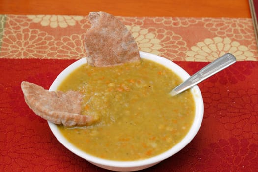 One bowl of split pea soup made with carrots. Two pieces of whole wheat pita bread and a spoon are dipped in split pea soup on a colorful place mat on a wooden table.