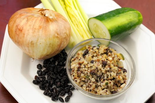 In a small round bowl is a cooked tricolor quinoa recipe surrounded by a bunch of black currants, a yellow onion, celery stalks and a half a cucumber on a plate resting on a wood surface..