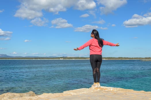 A woman stands on a rocky edge by the sea, arms open wide under a blue sky with scattered clouds, beautiful latina woman by the sea in Colonia San Jordi, Balearic Islands, Majorca, Spain,