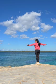 Woman standing near the ocean with arms open, enjoying the calm, dressed in pink and black, beautiful latina woman by the sea in Colonia San Jordi, Balearic Islands, Majorca, Spain