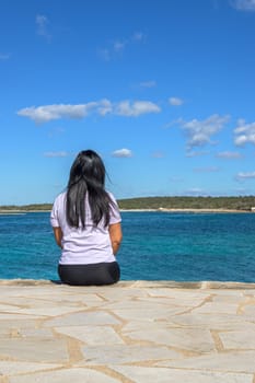 Latina woman, 48 years old, sitting on a pier in Colonia de Sant Jordi, Mallorca, Balearic Islands, Spain. A serene early spring day by the turquoise sea, basking in the sunshine and coastal breeze.