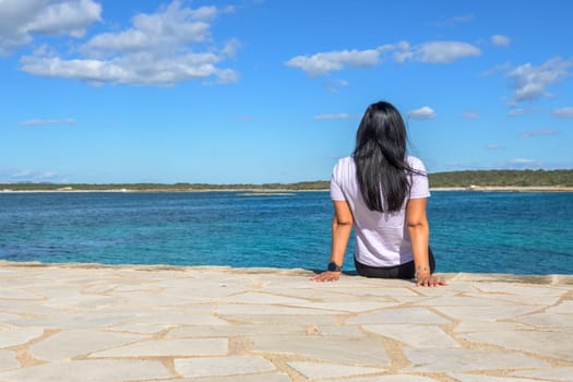 Rear view of a relaxed tourist sitting on a retaining wall while looking at the sea against the sky, in Colonia San Jordi, Balearic Islands, Majorca, Spain.
