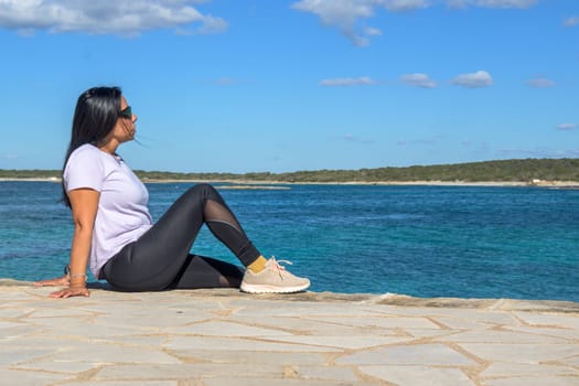 Woman sitting by the ocean, looking relaxed and peaceful, dressed casually on a sunny day, beautiful latina woman by the sea in Colonia San Jordi, Balearic Islands, Majorca, Spain