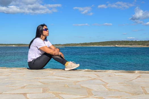 Latina woman, 48 years old, sitting on a pier in Colonia de Sant Jordi, Mallorca, Spain. A sunny early spring day by the turquoise sea, enjoying the peaceful coastal view.