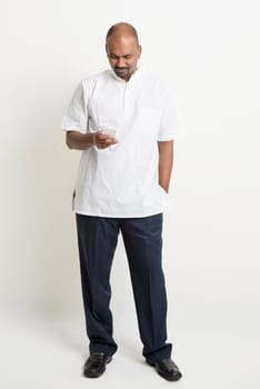 Portrait of full length mature casual business Indian man using smartphone, mobile apps concept, standing on plain background with shadow.