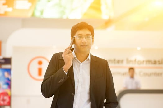 Asian Indian businessman walking by information counter, talking on phone, golden sunset background.