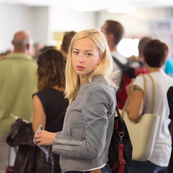 Young blond caucsian woman waiting in line with plain ticket in her hands. Lady standing in a long queue to board a plane.