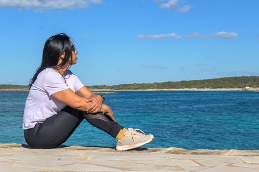 Latina woman, 48 years old, sitting on a pier in Colonia de Sant Jordi, Mallorca, Spain. A sunny early spring day by the turquoise sea, enjoying the peaceful coastal view.