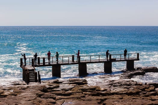 Fishermen on concrete pier jetty with blue ocean water along rocky coastline landscape.