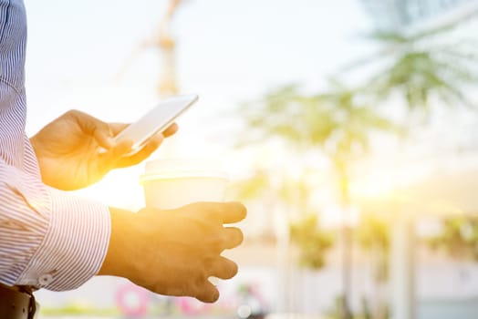 Man hand holding take away hot coffee cup and browsing smartphone in morning sunlight, outdoors business concept.