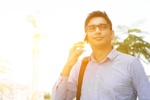 Indian businessman talking on phone in front modern office building. City view with beautiful sunlight.