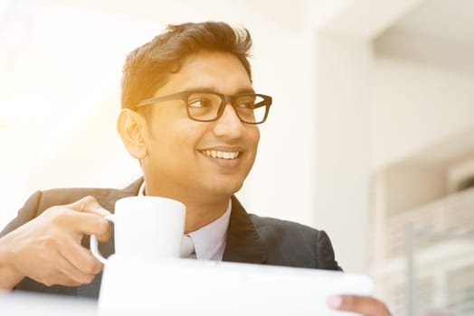 Young Asian Indian businessman using tablet pc at cafe, relaxing with a cup of coffee. India male business man, modern office building with sunlight as background.