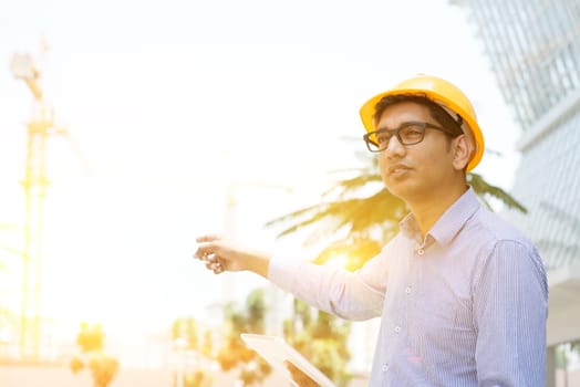 Portrait of a smiling Indian male contractor engineer with hard hat pointing to a crane at construction site
