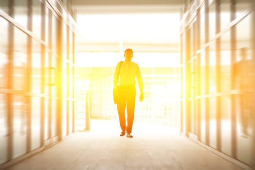 Asian Indian business man walking thru modern office building with glass window, bright backlight from sunset.